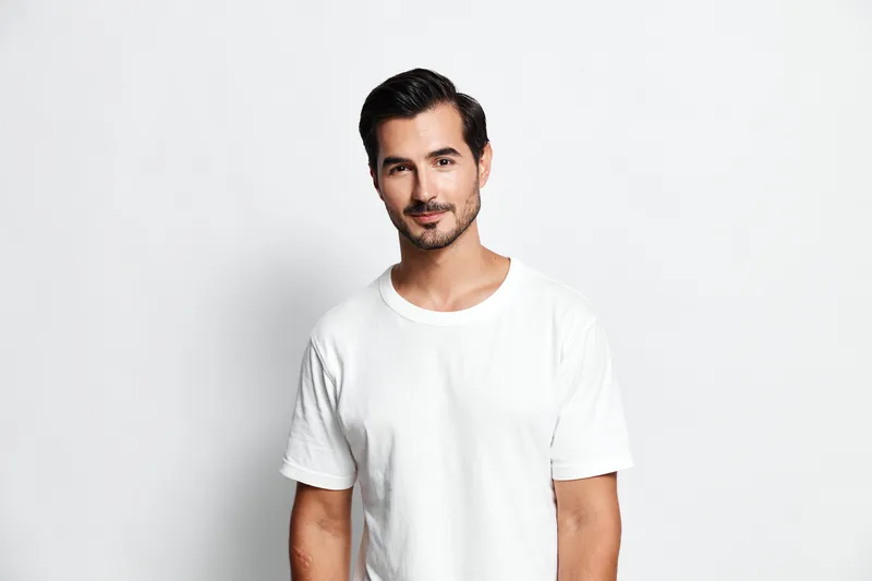 Charming smiling man with light brown hair wearing a casual white t-shirt poses confidently against a plain light background. Portrait lifestyle concept.