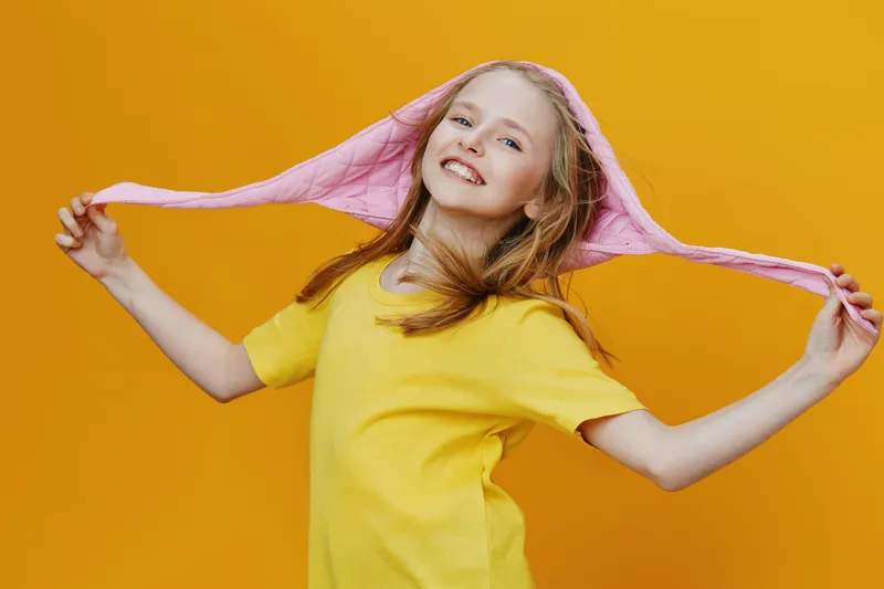 Cheerful girl in a yellow t shirt with a pink hoodie against a vibrant yellow background, expressing joy and playfulness