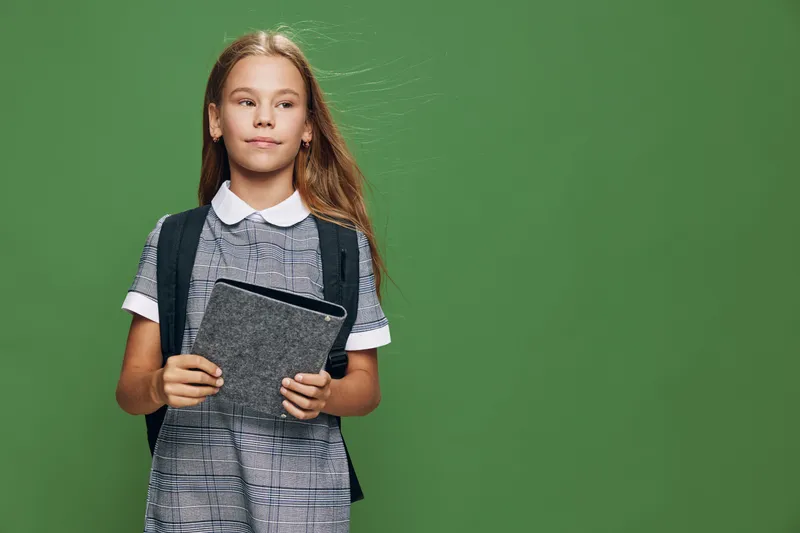 Girl student with long hair in school uniform and backpack holding tablet looking aside on green background. Education, technology and learning for child with digital device and childhood.
