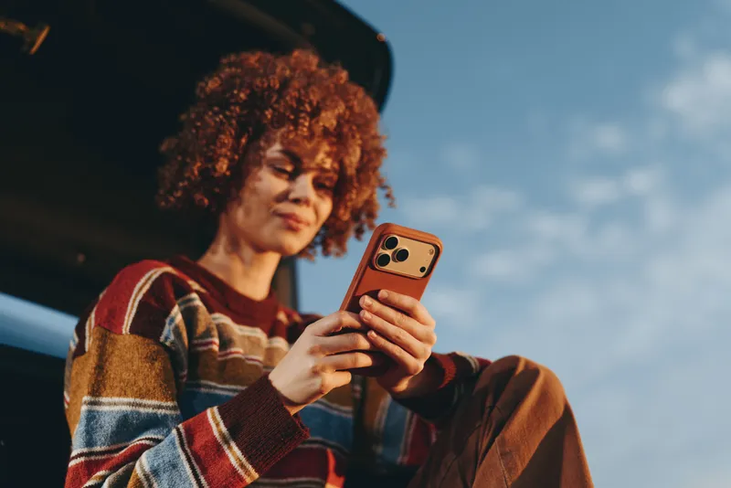 Woman with curly hair holding smartphone, portrait outdoors in a casual sweater, looking at screen. Daylight and blue sky background show a calm lifestyle moment today, naturally.