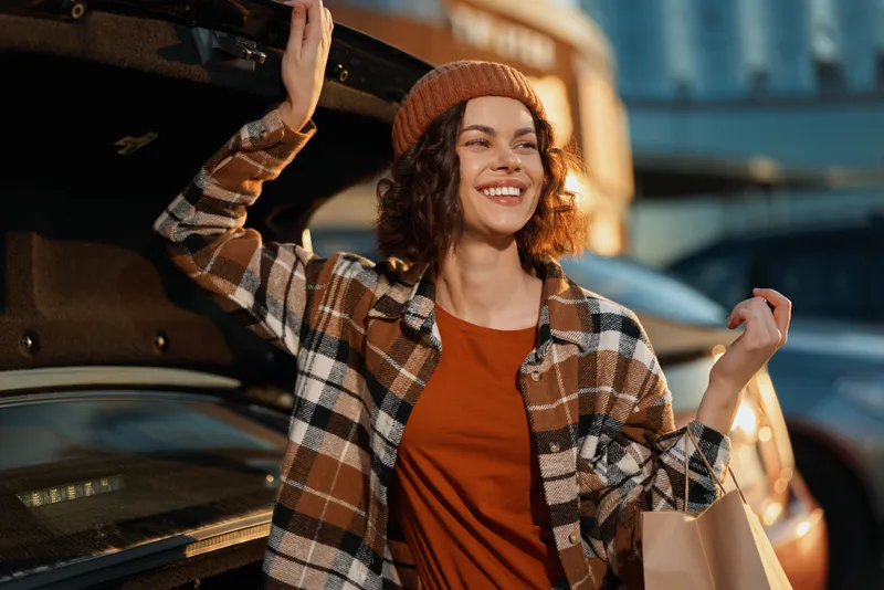 Woman smiling by car trunk with shopping bag, beanie and plaid jacket in urban parking, candid lifestyle and authenticity with golden hour glow, mindful living and emotional storytelling.