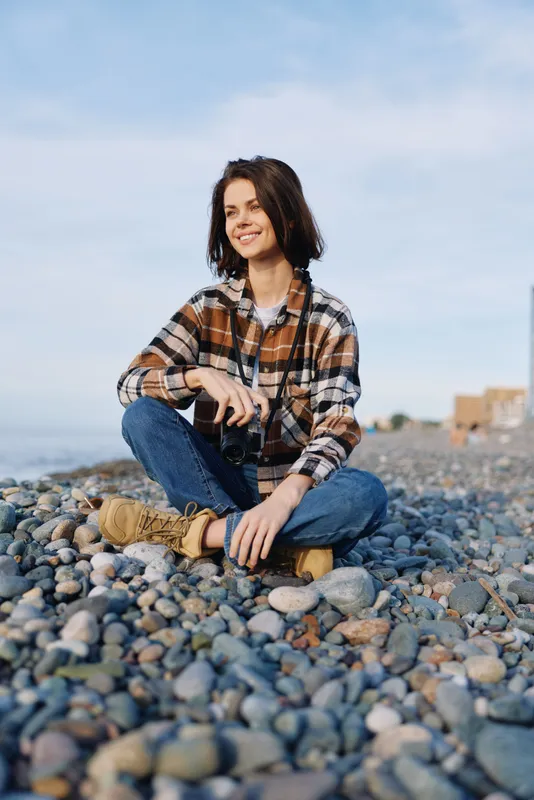 Smiling woman sits on a pebble beach with a camera hanging at her neck, wearing a plaid casual shirt and denim jeans, enjoying outdoor photography by the shore.