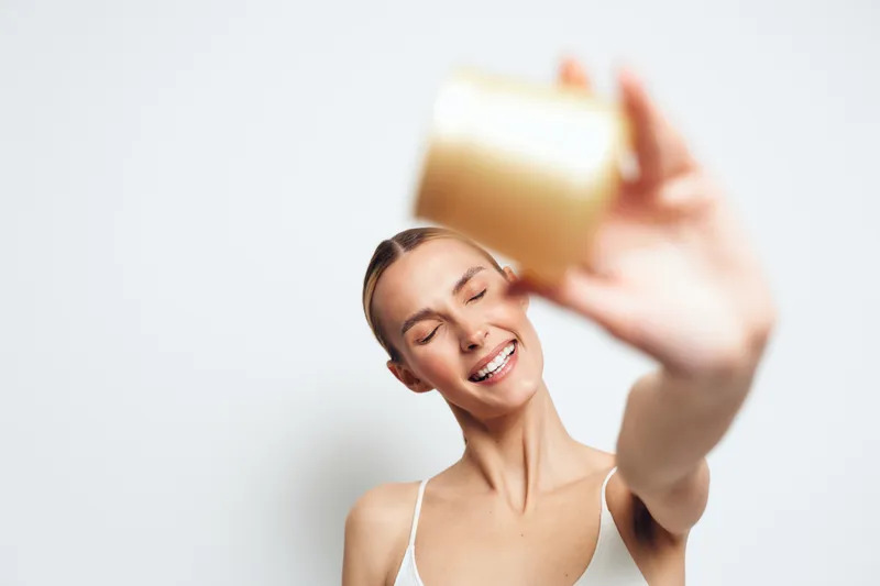 Cheerful woman indoors, smiling with eyes closed, reaching toward camera with bottle, soft lighting, white background, casual outfit, wellness moment, natural expression in bright mood