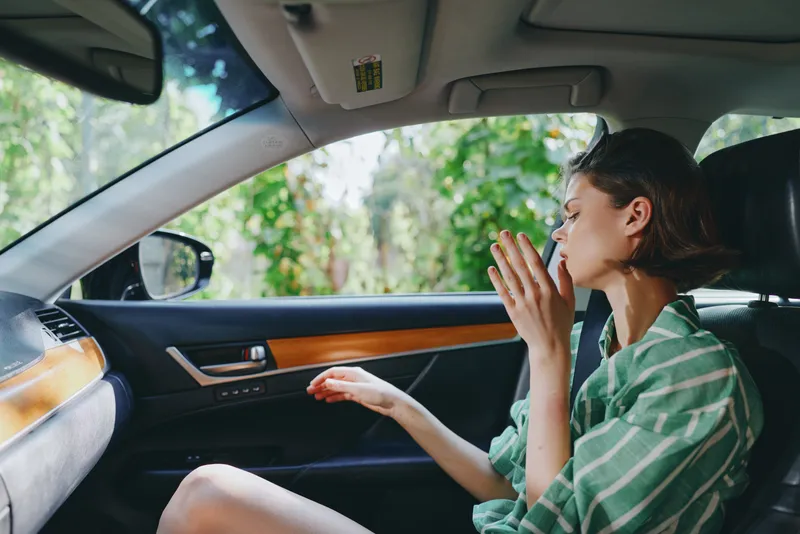 Woman sits in a parked car, eyes closed, hands raised in a contemplative gesture, sunlit interior and tranquil mood.