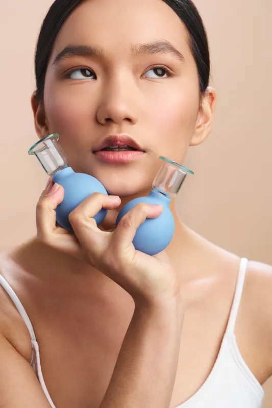 Asian woman holds two blue cosmetic bottles with clear caps near her chin, looking slightly upward with a calm, contemplative expression in a neutral studio setting, softly lit for a fresh skincare