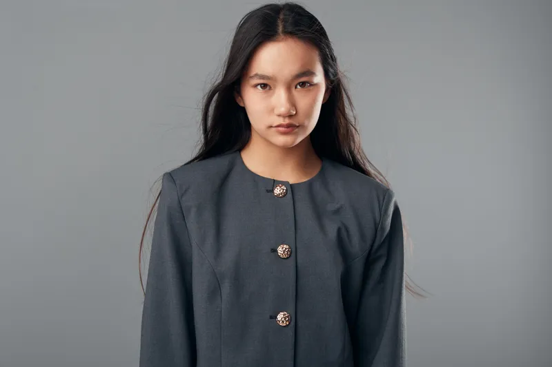 Young woman with long black hair wearing a gray blazer with decorative buttons looking forward on gray background. Studio portrait, serious expression and business casual style.