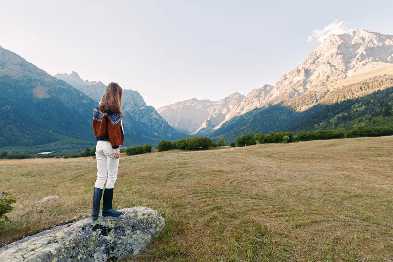 Woman with backpack standing on a rock in open field, hiking toward distant mountains and vast landscape, enjoying nature scenery and peaceful alpine vista at golden hour.
