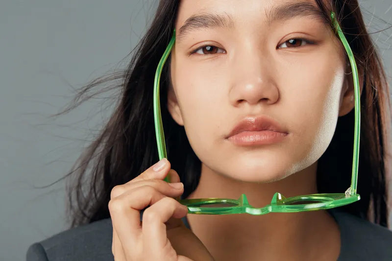 Young woman with long dark hair holds green glasses close to her face in closeup portrait. Serious expression with natural makeup and gray background in studio light.