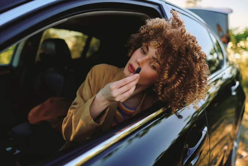 Smiling woman with curly hair applying lipstick in a car, lifestyle scene with retro film color, warm intimate tones, candid moment of beauty and wellbeing.
