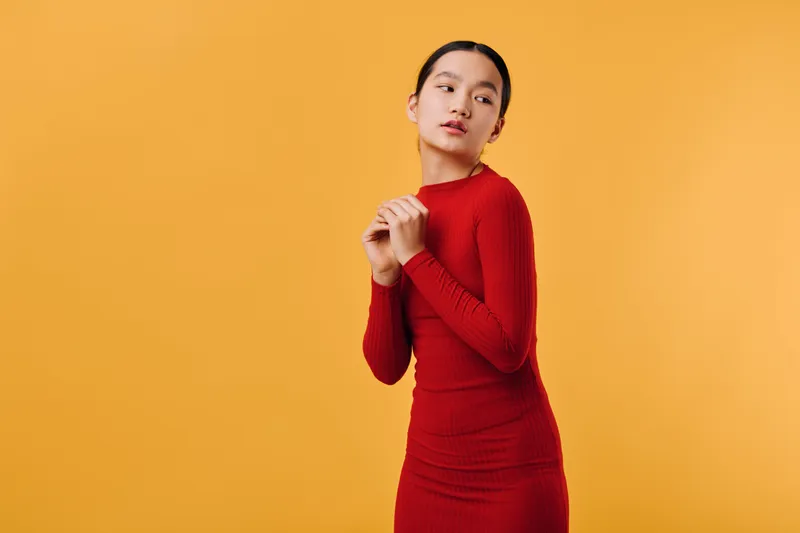 Woman in red dress posing on orange background, standing with hands close to face and looking sideways, fashion portrait with minimalist vibrant backdrop and confident expression.