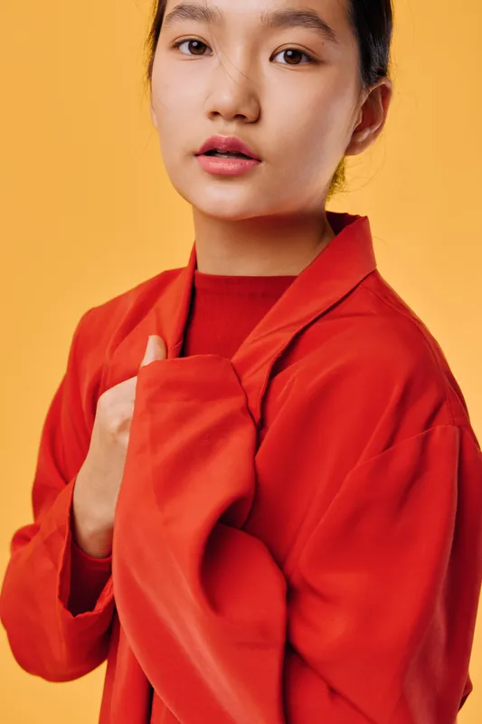 Young woman wearing red coat with hand clutching collar looking at camera with neutral expression on orange background in studio. Portrait fashion casual style and confident pose closeup.