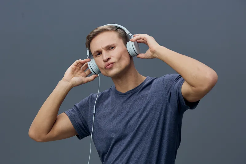 Young man with short hair listening to music through headphones, posing with a playful expression against a plain gray background. Studio portrait of a casual male enjoying music, showing personality 