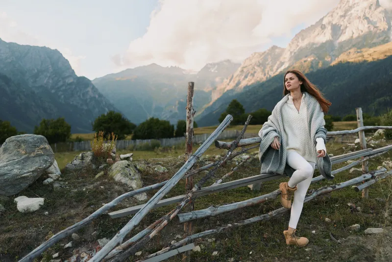 Casual woman in a cozy sweater posing by a wooden fence in a mountain landscape, capturing outdoors lifestyle and serene nature moments in soft daylight for a warm mood.