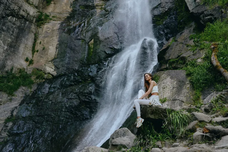 young woman sitting near waterfall in nature reserve, enjoying peaceful outdoor scenery, wearing white casual clothes, surrounded by rocks and greenery