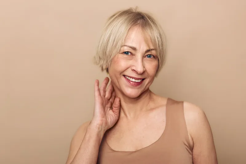 Smiling mature woman with short blonde hair touching neck, wearing beige tank top. Studio portrait on neutral background. Concept of natural beauty, aging, confidence, and healthy skin care.