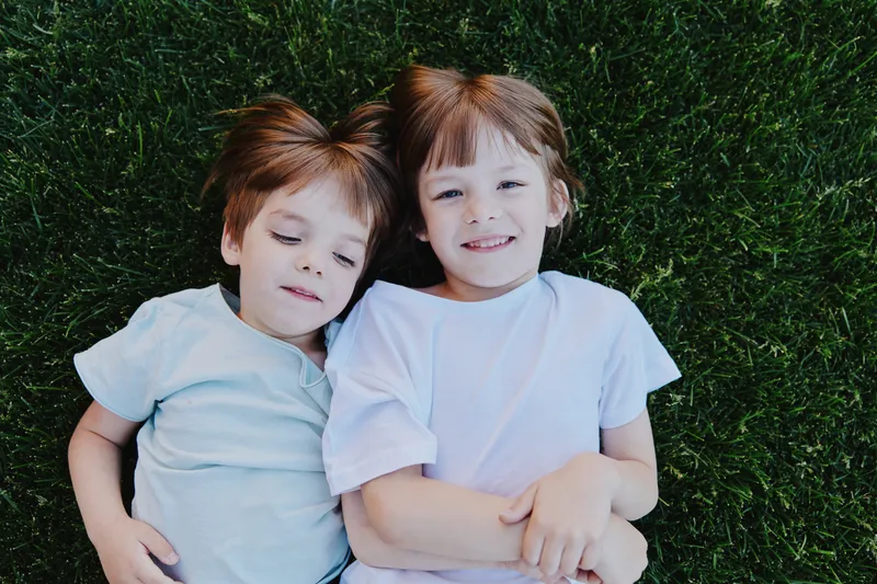 Two happy children lying on green grass, smiling joyfully while enjoying a sunny day outdoors. Their playful expressions radiate warmth and innocence.