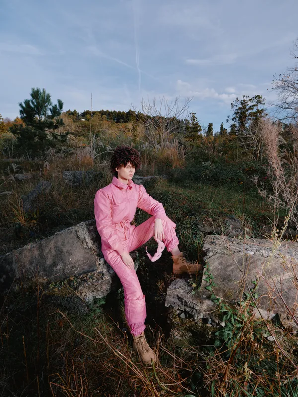 A fashion model sits on rugged rocks in a pink tracksuit, surrounded by autumnal foliage and open sky. This outdoor portrait highlights casual style, relaxed pose, and natural light in a rugged