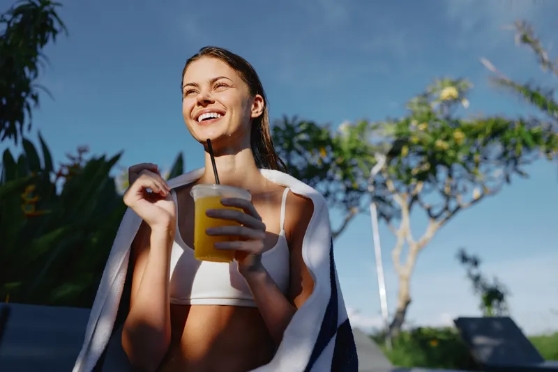 Happy woman outdoor yellow beverage smiling under bright sun, draped towel enjoying refreshing moment in garden scene, casual summer vibe, candid lifestyle moment captured for everyday wellness and