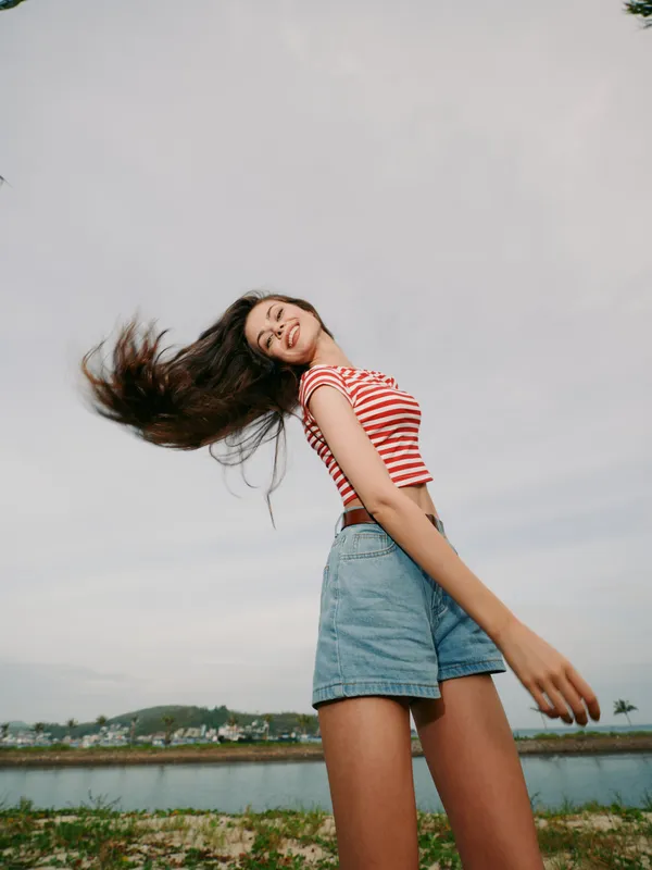 Young woman outdoors on a windy day wearing a striped crop top and denim shorts, standing beside a calm lake with distant hills, exuding carefree summer energy and movement.