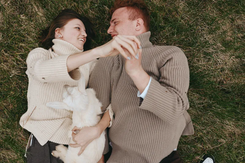 Carefree couple lying on grass with a goat kid, smiling and embracing, warm sweaters, candid outdoor scene, lifestyle portrait, rural charm, playful moment, soft natural light, cozy comfort,