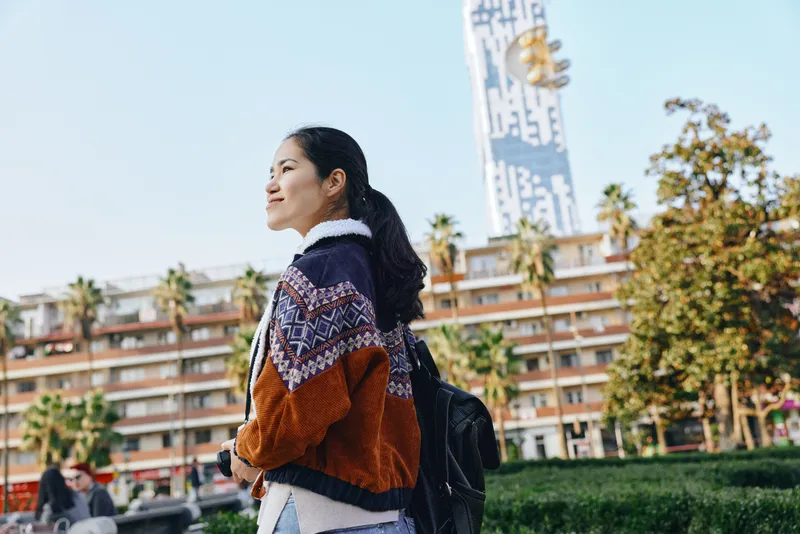 Young woman with backpack walks through a sunny urban square, wearing a patterned cardigan and jeans, exploring a cityscape with a tall clock tower and palm trees in the background.