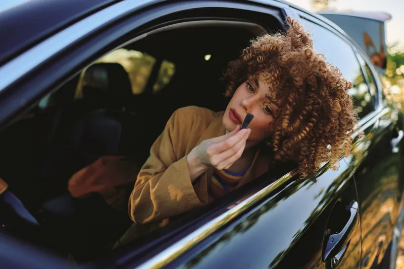 Woman applying makeup inside a sleek car, golden hour light, curly hair framing her face, casual wardrobe, retro film color mood, relaxed vibe