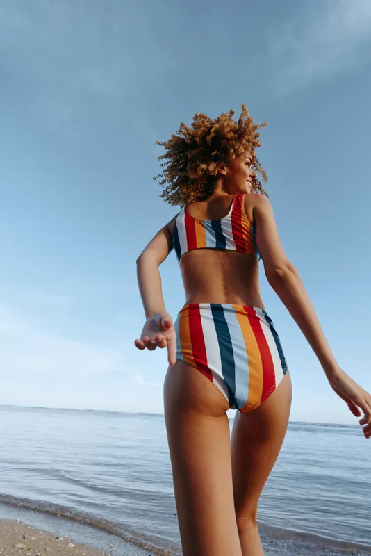 Woman in striped swimsuit walks along the beach by calm sea beneath blue sky, summer outdoors fashion model with relaxed arms extended, sunlit waves and sand, vertical photo portrait