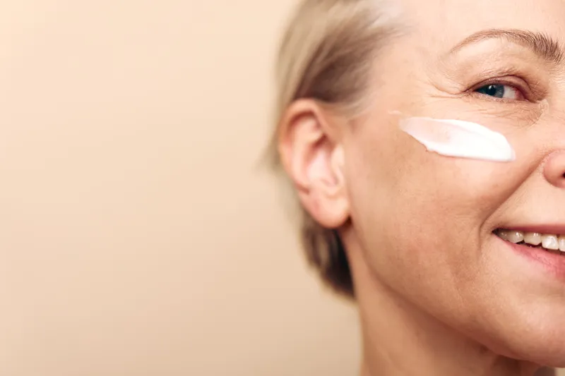 Close-up of happy mature woman with short hair applying white face cream on cheek. Skincare concept with natural beauty, wellness, healthy skin, and anti-aging care on beige background.