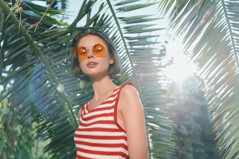 A cheerful woman in a red and white striped sleeveless top and round sunglasses stands in a sunny tropical setting, looking up with a smile, creating a bright, carefree vacation mood.