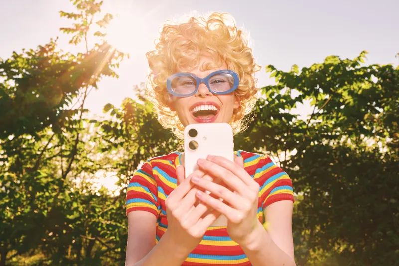 A young woman with curly blonde hair wearing a colorful striped shirt and blue glasses laughs while taking a selfie outdoors on a sunlit day.
