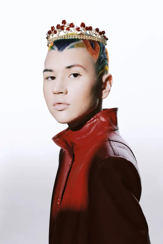 Portrait of a woman with colorful hair and a decorative crown wearing a red jacket, captured against a clean white background with soft lighting and sharp details.