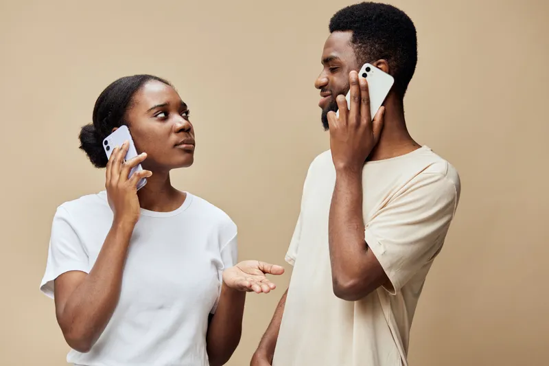 Young African couple talking on smartphones, expressing emotions with casual clothing, warm beige background, modern lifestyle, communication concept
