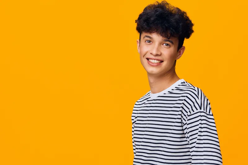 smiling young man with curly hair wearing striped shirt stands against bright orange background, expressing happiness and youthful energy in casual style.