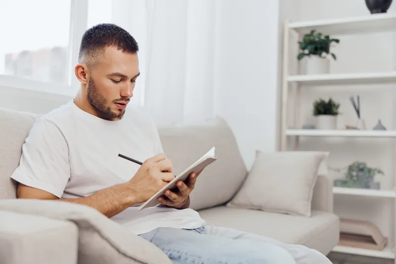 Thoughtful man writing in a notebook at home, surrounded by natural light and minimalistic decor, reflecting creative ideas and inspiration for projects.