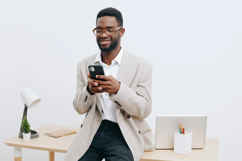 Smiling man using smartphone in modern office environment, casual business attire, cheerful expression, workspace setup, bright interior, professional atmosphere, communication concept, lifestyle phot