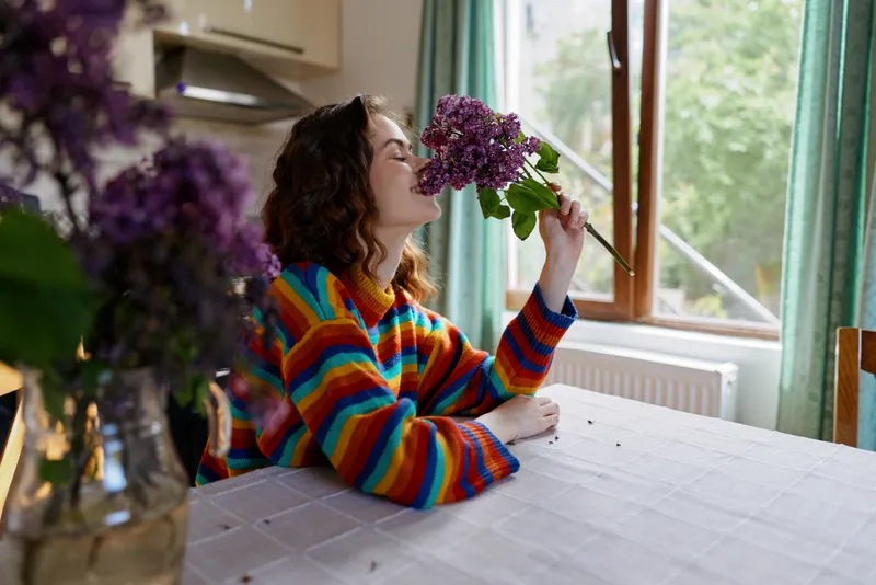Woman wearing a colorful striped sweater sits at a wooden table indoors, smelling a bouquet of purple lilacs near the window, conveying a warm home lifestyle, cozy creative mood, and gentle everyday
