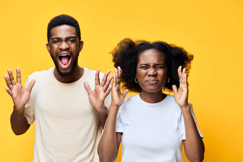Young Black couple expressing strong emotions with yelling and frustrated gestures against a bright yellow background, showcasing joy and distress in vibrant attire