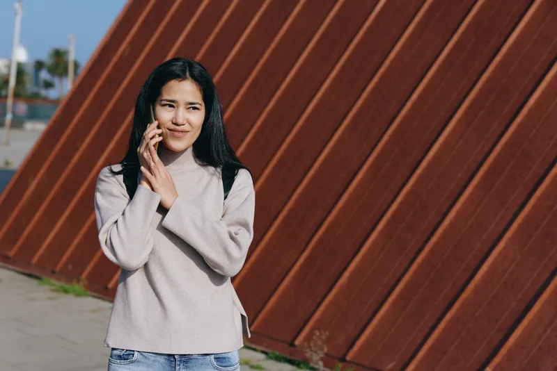 Woman talking on a mobile phone outdoors beside a diagonal wooden wall, wearing a light knit sweater and denim jeans, casual street scene, daytime light, candid moment of everyday communication