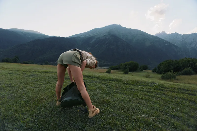 Woman outdoors in a grassy field near mountains, bending to pick up a backpack, casual lifestyle shot, natural lighting, sense of exploration and calm.
