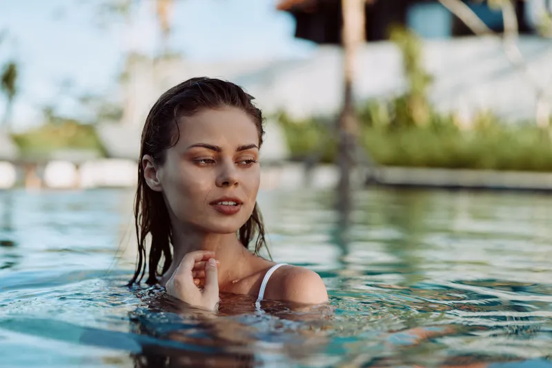 A woman in swimsuit stands in an outdoor pool, enjoying leisure and relaxation under bright sun, with water reflections and tropical scenery in the background, conveying a carefree vacation mood.