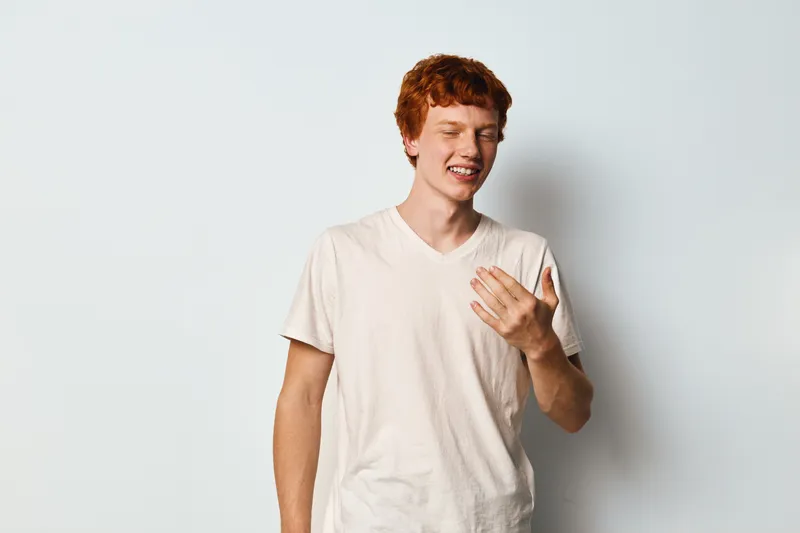 Young man with curly red hair wearing white t shirt is smiling with closed eyes and candid laughter on light background. Casual portrait of happy male emotion and gesture.