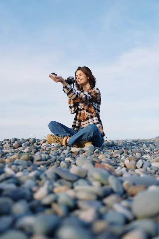 Outdoor woman sitting on pebbles, camera in hand, joyful moment under blue sky, casual lifestyle shot for travel or nature photography.