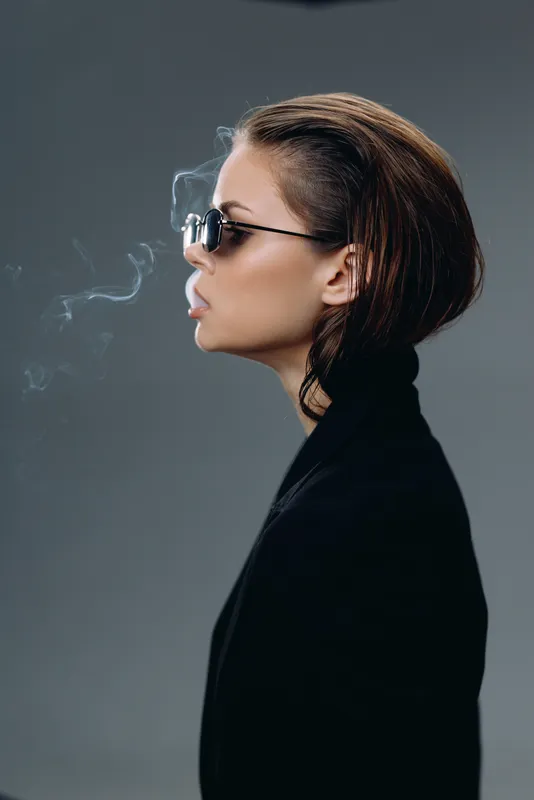 Woman wearing sunglasses smoking cigarette with smoke against dark background in stylish outfit