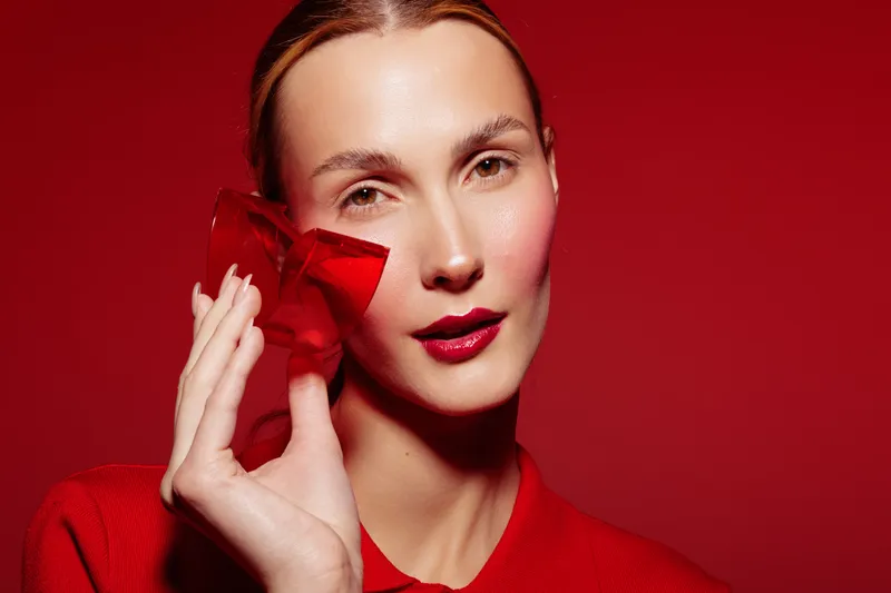 Young woman with clear skin holds red translucent object near face on red background. Concept of beauty, skincare, fashion, and modern minimalism with vibrant color contrast and soft lighting.
