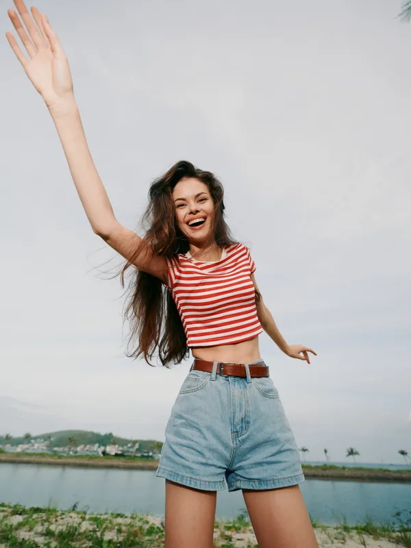 Young woman enjoying sunny day outdoors, raising one arm in excitement, smiling brightly, wearing striped crop top and denim shorts, carefree summer vibe.