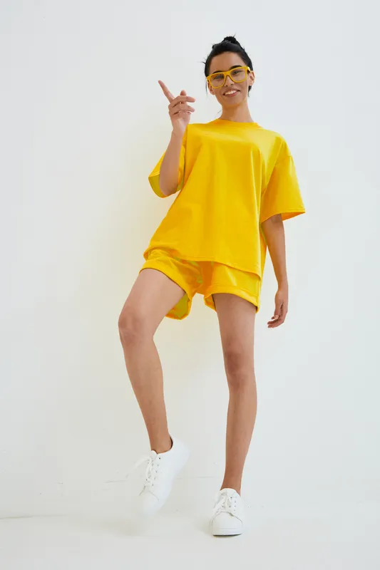 smiling woman in bright yellow casual outfit and sunglasses standing against white background, wearing shorts and white sneakers, cheerful and relaxed mood