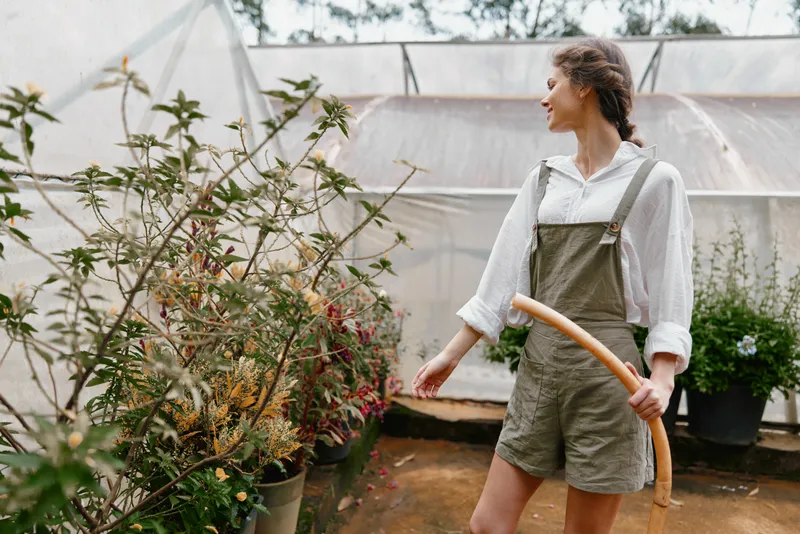 Young woman in a greenhouse wearing a white shirt and green overalls, tending to plants with a hose, expressing joy and connection to nature