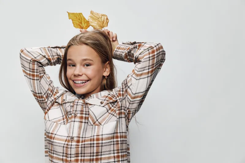 Girl smiling and posing with autumn leaves on head wearing plaid shirt, casual style with hands behind head against white background. Happy child with fall season theme and natural expression.