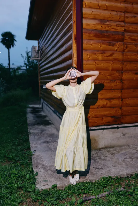A woman in a yellow dress stands beside a wooden building at dusk, covering her face with her hands, creating a playful mysterious mood in an outdoor rural setting.