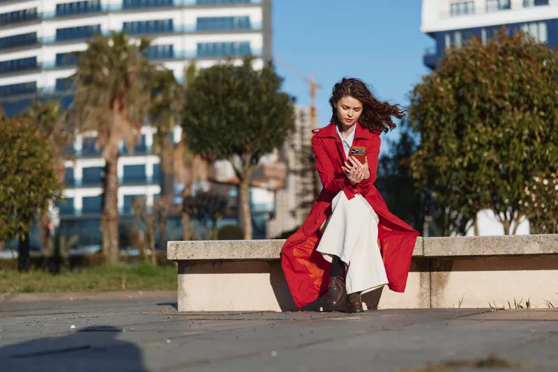 A woman sits on a bench in an urban outdoor setting wearing a red coat, posing in a portrait while checking a phone, showcasing fashion and modern style against a bright daytime street scene